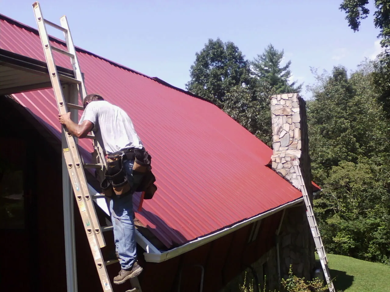 Roofer on a ladder beside a red metal roof and stone chimney — Soffit & Fascia Repair by Roofing Brothers in Holmes